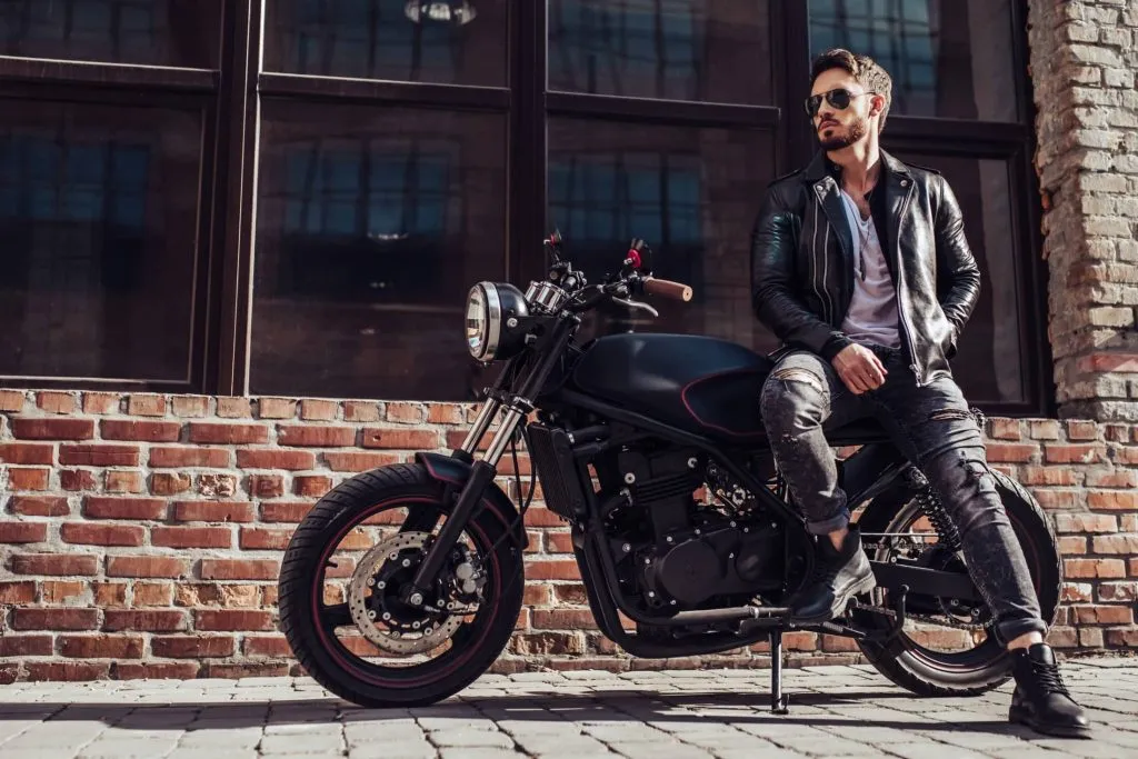 Mann mit schwarzer Lederjacke und Sonnenbrille sitzt seitwärts auf einem Motorrad