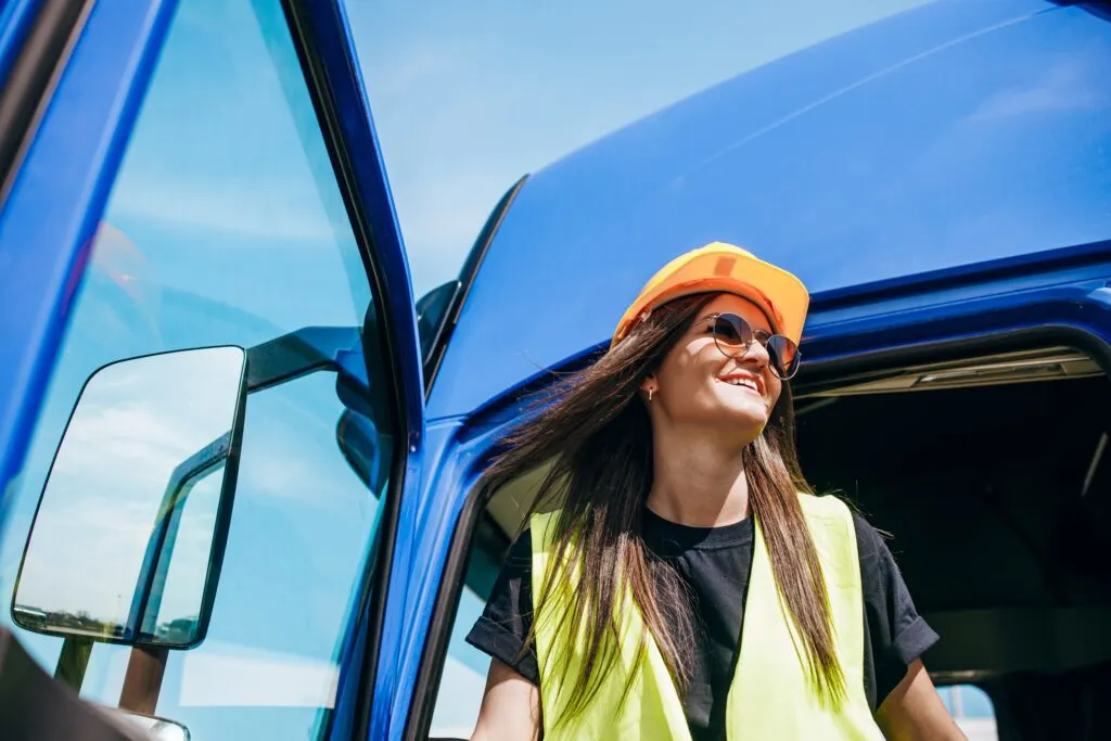Frau trägt einen Orangen Helm und eine Sonnebrille und steigt aus einem blauen LKW aus
