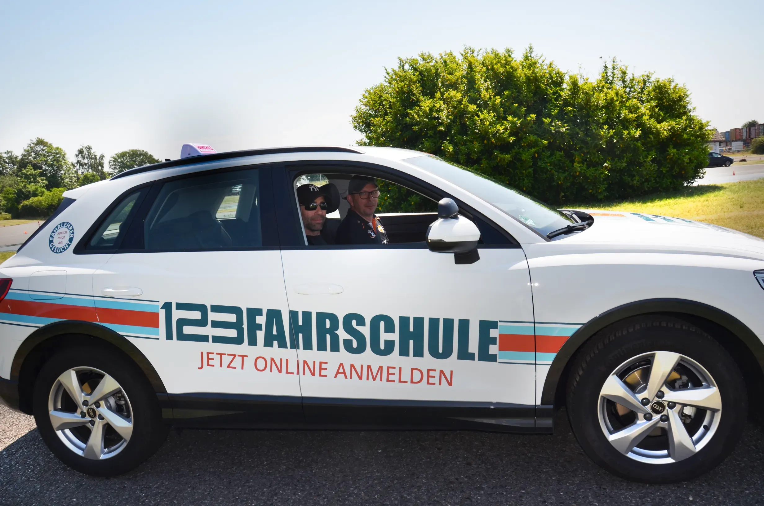In einem Auto mit der Werbeauschrift der 123Fahrschule vor einer grünen Landschaft sitzen zwei Fahrlehrer und lächeln in die Kamera