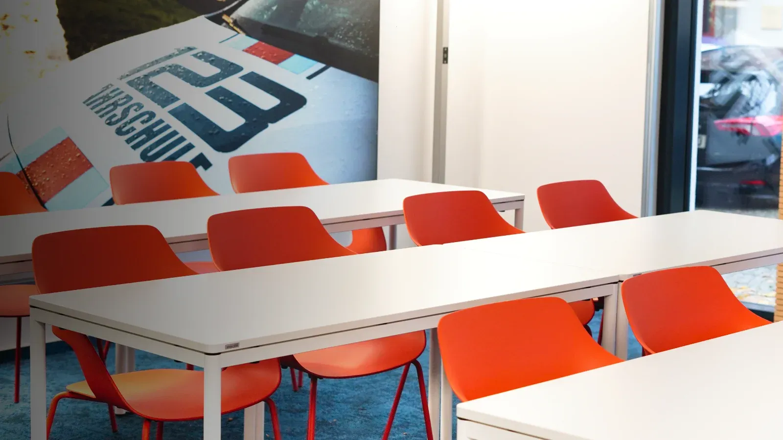 Ein Schlungsraum der 123fahrschule mit orangenen Stühlen und weißen Tischen. Im Hintergrund befindet sich ein Bild mit einem Auto der 123fahrschule drauf.
